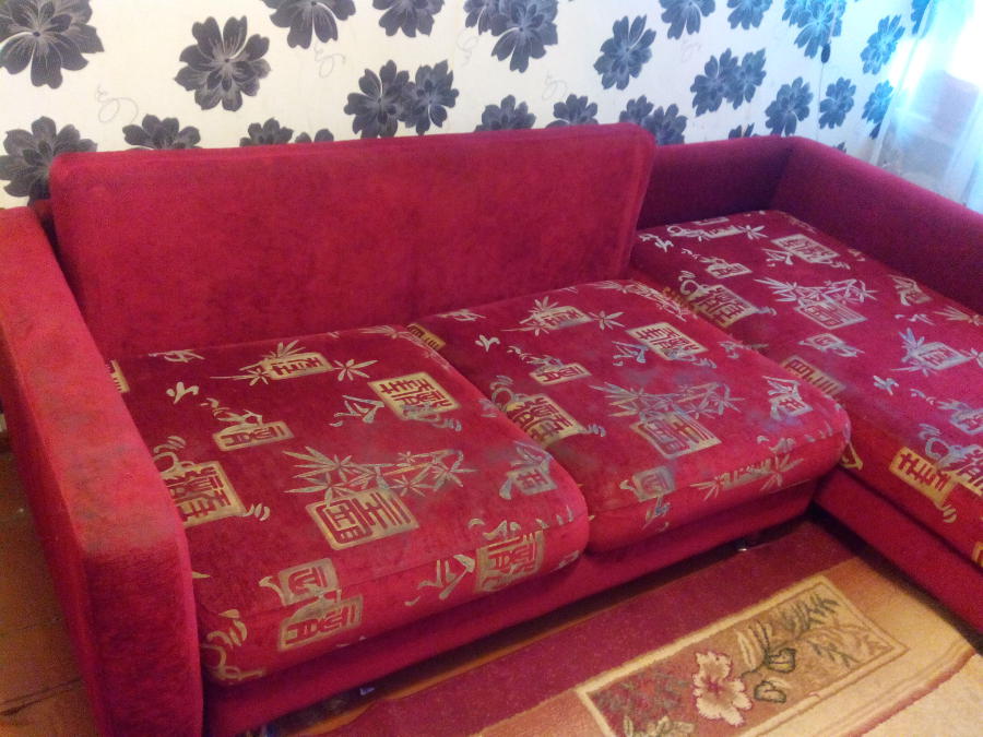 Фото красного углового дивана до чистки, видим различные загрязнения и засаленные места.